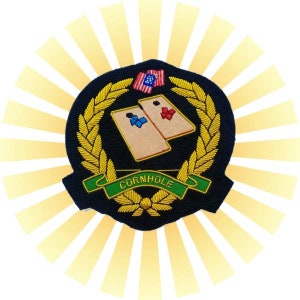 May include: A navy blue embroidered patch with a gold wreath, two beige cornhole boards, a green ribbon with the text "CORNHOLE", and a small American flag.
