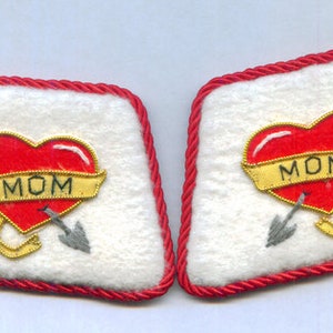 May include: Two white felt patches with red borders, each featuring a red heart with a gold ribbon that says "MOM" and two silver arrows piercing the heart.