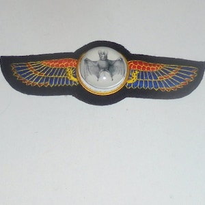 May include: A black fabric patch with a red, blue, and gold embroidered design of wings. A round glass dome with a white background and a black and white image of a bat is centered on the patch.