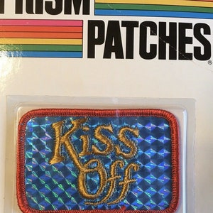 May include: A rectangular patch with the words "Kiss Off" embroidered in gold thread on a blue, iridescent background. The patch has a red border and is packaged on a white card with the words "PRISM PATCHES" in black.