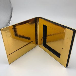 May include: A gold-colored box with a black border and a cutout in the center. The box is open and the inside is visible.