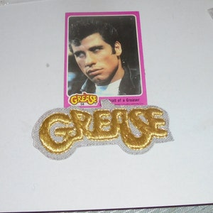 May include: A gold and silver embroidered patch with the word "Grease" in a stylized font. The patch is next to a trading card featuring a portrait of an actor from the movie Grease.