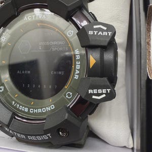 May include: A black and olive green digital wristwatch featuring a round face with the text ACTIVA, 1/100S CHRONO, SPORTS, ALARM, CHIME, and WR 3BAR. The side includes prominent black buttons labeled START and RESET with white text and arrows.