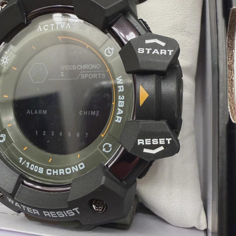 May include: A black and olive green digital wristwatch featuring a round face with the text ACTIVA, 1/100S CHRONO, SPORTS, ALARM, CHIME, and WR 3BAR. The side includes prominent black buttons labeled START and RESET with white text and arrows.
