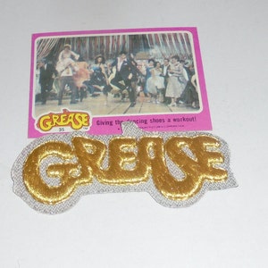 May include: A gold-colored "Grease" patch with a white background. The patch is a promotional item for the movie "Grease".