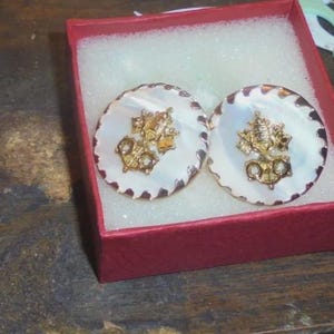 May include: Pair of round, gold-toned cufflinks with a nautical design, featuring a gold anchor and other maritime symbols. The cufflinks have a white, iridescent mother-of-pearl center and a scalloped edge. Presented in a red gift box with white padding.