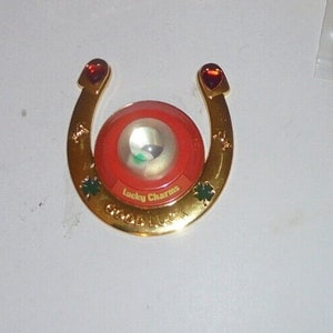 May include: Gold horseshoe charm with a red and white Lucky Charms cereal box design. The horseshoe has a small green shamrock on each side and the words "Good Luck" are inscribed on the bottom.