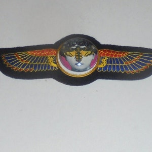 May include: A black fabric patch with gold, red, and blue embroidered wings. A round glass dome with a white and pink image of a face with a bee inside is centered on the patch.