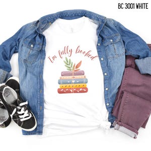 May include: White t-shirt with the text "I'm fully booked" and a stack of colorful books. The shirt is paired with a denim jacket, burgundy pants, and black sneakers. The shirt's design features a stack of books tied with a string and leaves.