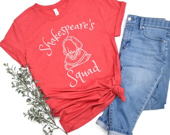 May include: A heather red t-shirt with the text "Shakespeare's Squad" and a graphic of William Shakespeare. The shirt is paired with light blue denim jeans. A sprig of greenery is on the left side of the image.