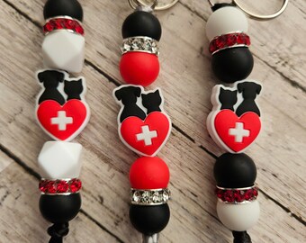 May include: Three keychains with black, white, and red beads. Each keychain features a charm of two dogs with a red heart and a white cross. The keychains have different colored tassels.