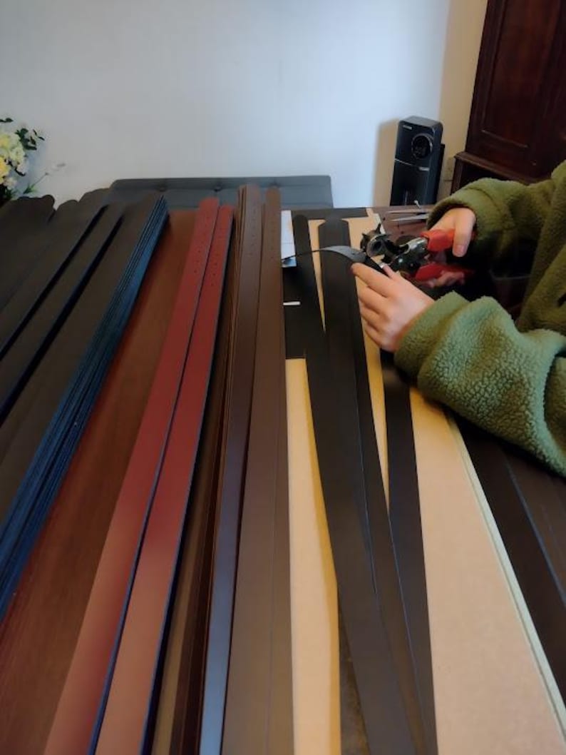 Puede incluir: Varias tiras de cuero en diversos colores, incluyendo negro, marr&oacute;n y burdeos, dispuestas sobre una mesa. Una persona est&aacute; usando una herramienta para trabajar el cuero. Las tiras parecen ser para proyectos de artesan&iacute;a o marroquiner&iacute;a.