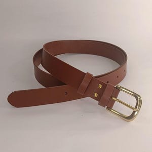 May include: A smooth brown belt featuring a polished gold-tone metal buckle and two matching leather keepers. The belt strap has a rounded tip and includes two gold-tone rivets securing the buckle assembly to the main body of the belt.