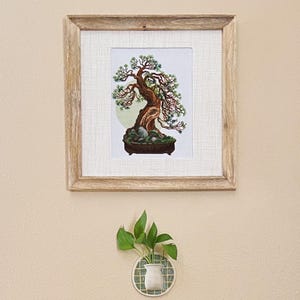 May include: A framed cross-stitch of a bonsai tree with a light wood frame. The tree has a brown trunk, green foliage, and sits in a brown pot with rocks. Below, a small white vase with green leaves is mounted on a wall.