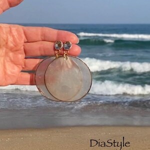 May include: A pair of large, circular earrings with a gold-colored frame and a pearlescent, translucent center. The earrings are held up against a backdrop of the ocean and beach. The earrings have a small, clear gem at the top. The text "DiaStyle" is visible.