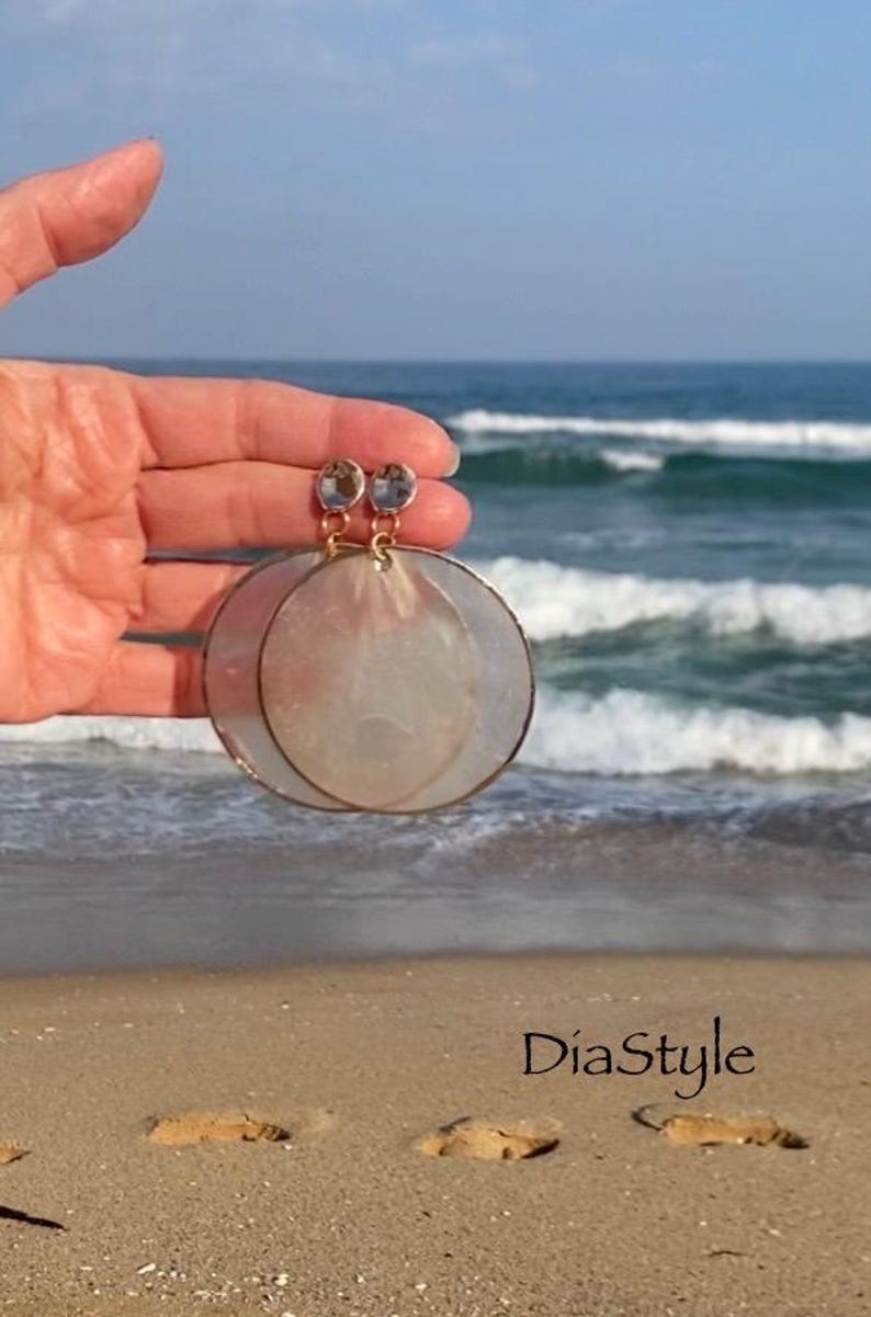 May include: A pair of large, circular earrings with a gold-colored frame and a pearlescent, translucent center. The earrings are held up against a backdrop of the ocean and beach. The earrings have a small, clear gem at the top. The text "DiaStyle" is visible.