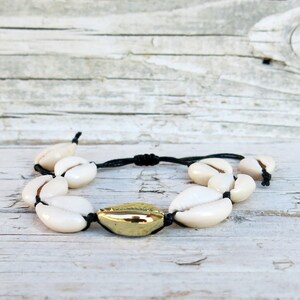 K&ouml;nnte beinhalten: Ein Armband aus wei&szlig;en Muscheln mit einer goldenen Akzentperle. Das Armband ist aus schwarzem Band gefertigt und mit einem Knoten gebunden.