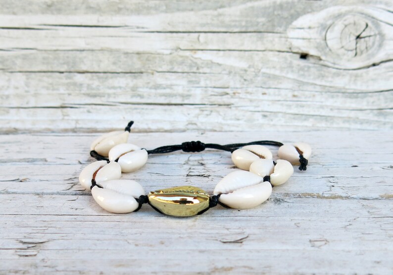 K&ouml;nnte beinhalten: Ein Armband aus wei&szlig;en Muscheln mit einer goldenen Akzentperle. Das Armband ist aus schwarzem Band gefertigt und mit einem Knoten gebunden.