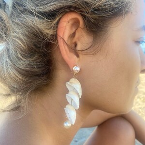 May include: White shell earrings with a pearl accent. The earrings are long and dangle from the earlobe.