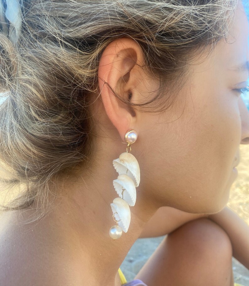 May include: White shell earrings with a pearl accent. The earrings are long and dangle from the earlobe.