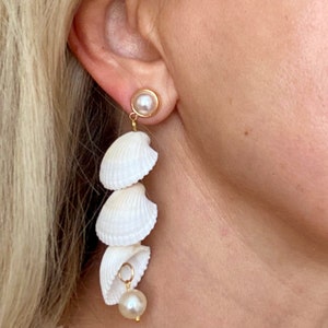 May include: A pair of long, white shell earrings with a pearl dangle. The earrings are made of natural seashells and are a beautiful and unique piece of jewellery.