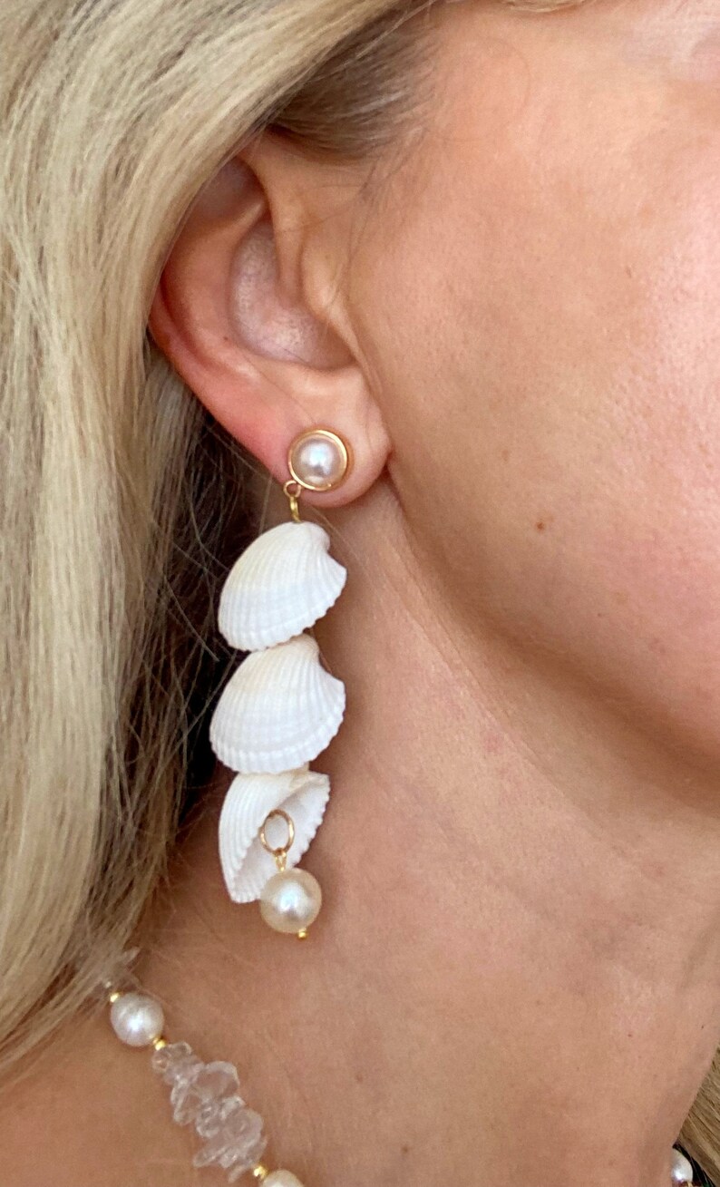 May include: A pair of long, white shell earrings with a pearl dangle. The earrings are made of natural seashells and are a beautiful and unique piece of jewellery.
