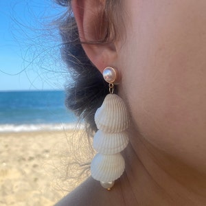 May include: A pair of long, white seashell earrings with a pearl accent. The earrings are made of natural seashells and are perfect for a beachy look.