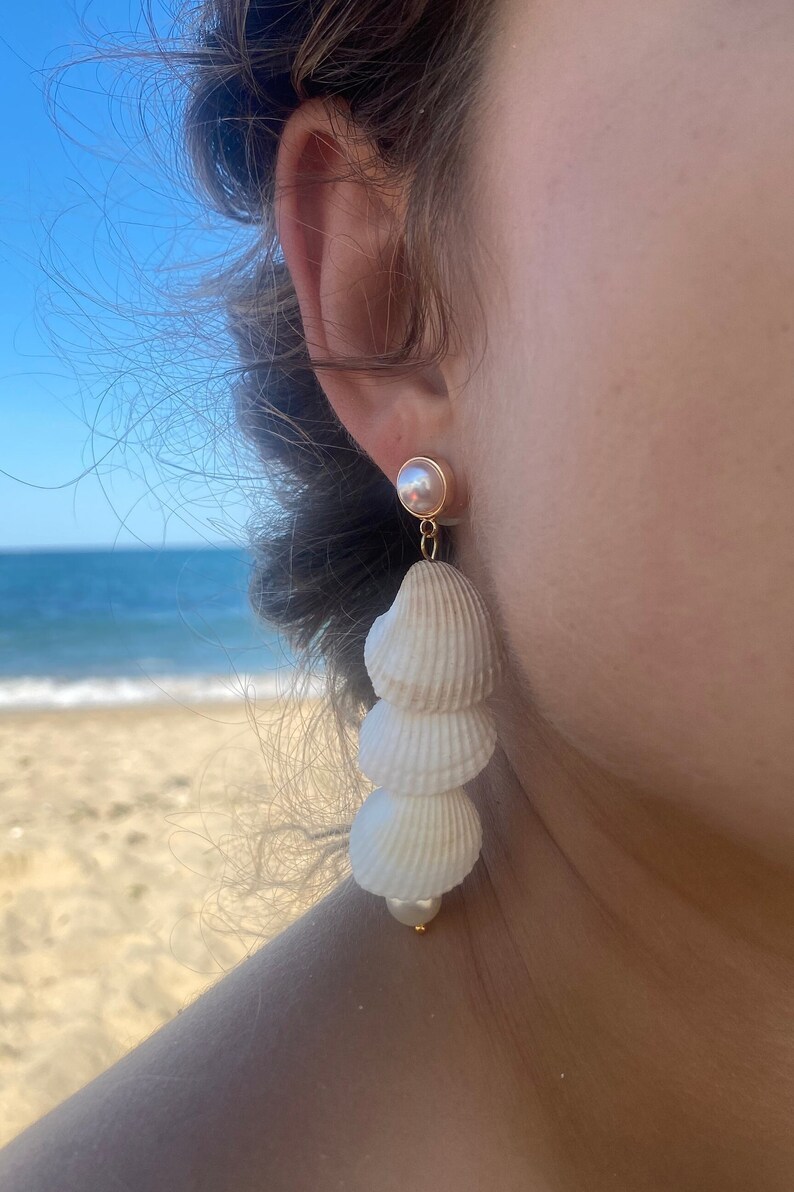 May include: A pair of long, white seashell earrings with a pearl accent. The earrings are made of natural seashells and are perfect for a beachy look.