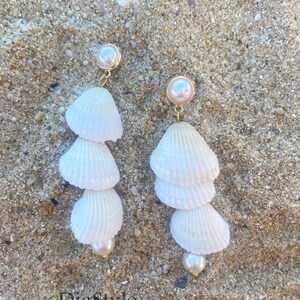 May include: A pair of white seashell earrings with gold accents and pearl accents. The earrings are hanging against a sandy background.