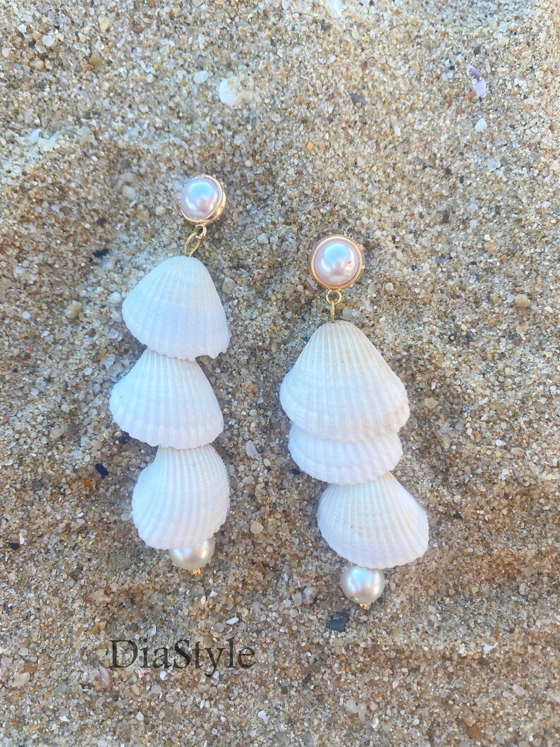 May include: A pair of white seashell earrings with gold accents and pearl accents. The earrings are hanging against a sandy background.