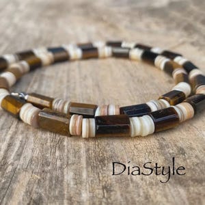 Puede incluir: Un collar de cuentas que alterna cuentas rectangulares de piedra ojo de tigre con pequeñas cuentas planas y circulares de color blanco y beige. El collar está dispuesto en forma circular e incluye un cierre de metal de tono plateado en el extremo.