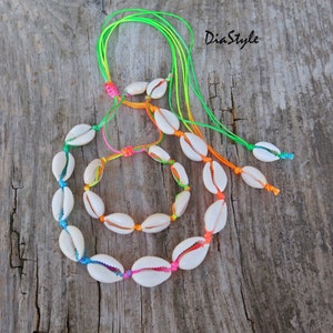 Op de afbeelding: Een set van twee kettingen gemaakt met witte schelpen en kleurrijk touw. De langere ketting heeft een regenboog aan kleuren, terwijl de kortere ketting een groen en oranje touw heeft.