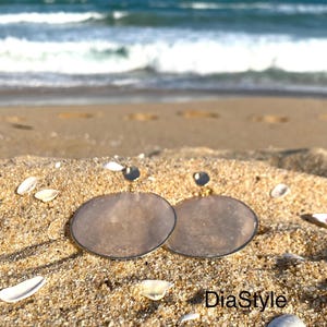 May include: Two pairs of silver-toned, circular earrings with a translucent, light brown center, resting on sand with seashells. The earrings are set against a backdrop of the ocean and a blue sky. The text "DiaStyle" is visible.