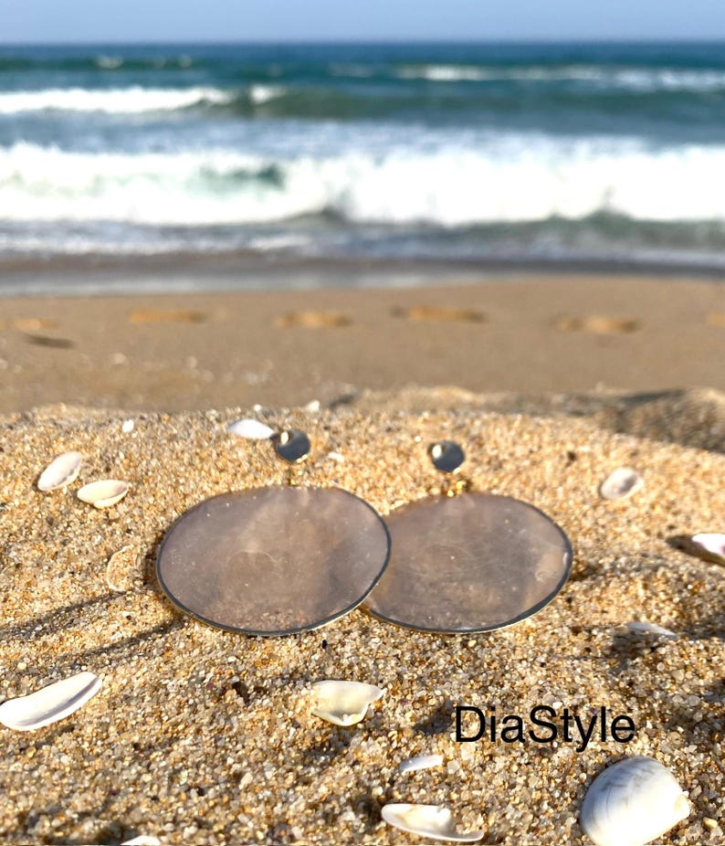 May include: Two pairs of silver-toned, circular earrings with a translucent, light brown center, resting on sand with seashells. The earrings are set against a backdrop of the ocean and a blue sky. The text "DiaStyle" is visible.