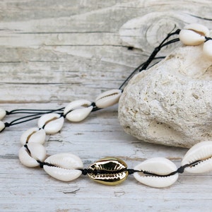 K&ouml;nnte beinhalten: Eine wei&szlig;e Muschelkette mit goldener Verzierung. Die Kette besteht aus Kaurischnecken, die mit schwarzem Band zusammengef&uuml;gt sind.