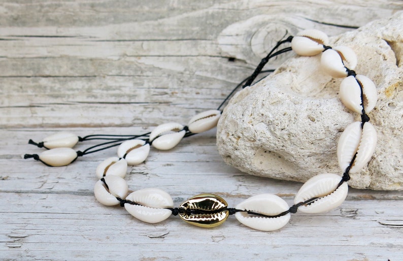 K&ouml;nnte beinhalten: Eine wei&szlig;e Muschelkette mit goldener Verzierung. Die Kette besteht aus Kaurischnecken, die mit schwarzem Band zusammengef&uuml;gt sind.