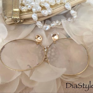 May include: A pair of large, gold-toned hoop earrings with a pearlescent, oval-shaped center. The earrings are displayed on a bed of white fabric flowers, with a gold clutch and pearl necklace in the background. The text "DiaStyle" is visible.