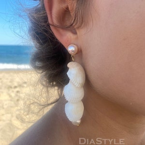 May include: A pair of long, white shell earrings with a pearl accent. The earrings are dangling from the earlobe and have a gold hook.