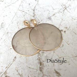 May include: A pair of large, round earrings with a gold-colored trim and posts. The earrings have a light, pearlescent center. The earrings are displayed on a textured, light-colored surface. The text "DiaStyle" is visible.