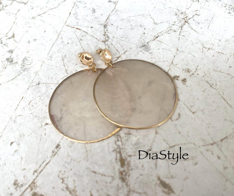 May include: A pair of large, round earrings with a gold-colored trim and posts. The earrings have a light, pearlescent center. The earrings are displayed on a textured, light-colored surface. The text "DiaStyle" is visible.