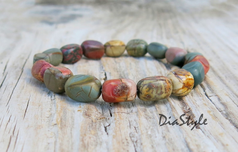 May include: A bracelet made of colorful, oval-shaped stones. The stones are a mix of brown, green, red, and white. The bracelet is on a wooden surface.