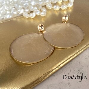 May include: Gold-toned, circular earrings with a textured, light-colored center. The earrings are set against a gold clutch, with a string of pearls in the background. The earrings have a post backing. The text "DiaStyle" is visible.