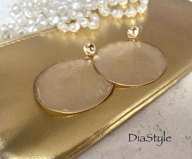 May include: Gold-toned, circular earrings with a textured, light-colored center. The earrings are set against a gold clutch, with a string of pearls in the background. The earrings have a post backing. The text "DiaStyle" is visible.
