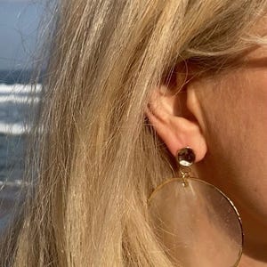 May include: Close-up of a person wearing large, gold-rimmed earrings with a circular, translucent design. The earrings are attached to a small, dark-colored stud. The text "DiaStyle" is visible in the lower left corner.