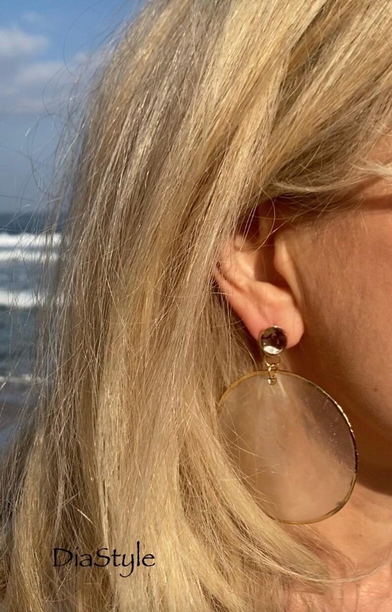 May include: Close-up of a person wearing large, gold-rimmed earrings with a circular, translucent design. The earrings are attached to a small, dark-colored stud. The text "DiaStyle" is visible in the lower left corner.