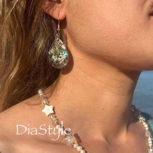 Pode incluir: Um brinco em forma de lágrima com um mosaico de fragmentos de concha de abalone iridescente em tons de verde, azul e prata. Um colar com contas brancas em forma de estrela, contas de cristal transparente e pequenas pérolas com o texto DiaStyle.