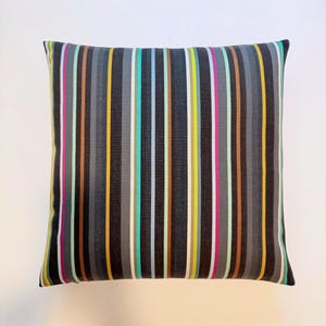 May include: A square throw pillow featuring a vertical striped pattern in shades of charcoal gray, black, mustard yellow, light blue, mint green, and hot pink. The fabric has a visible woven texture with consistent, repeating lines across the surface.