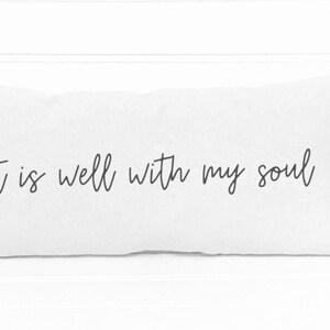 May include: White rectangular pillow with the phrase "It is well with my soul" in black script. The pillow is made of a soft, textured fabric and is a decorative item for a bed or couch.