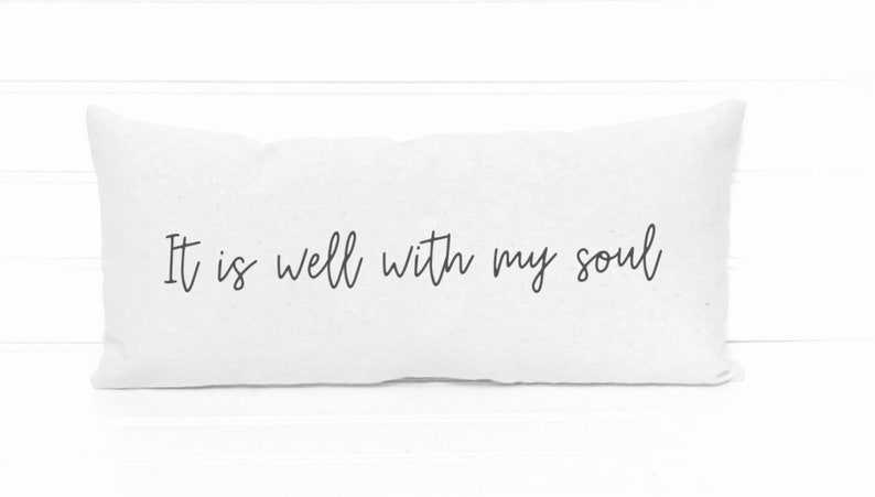May include: White rectangular pillow with the phrase "It is well with my soul" in black script. The pillow is made of a soft, textured fabric and is a decorative item for a bed or couch.