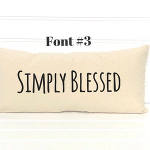 May include: A white pillow with the text "SIMPLY BLESSED" in black font. The pillow is on a white wood background.